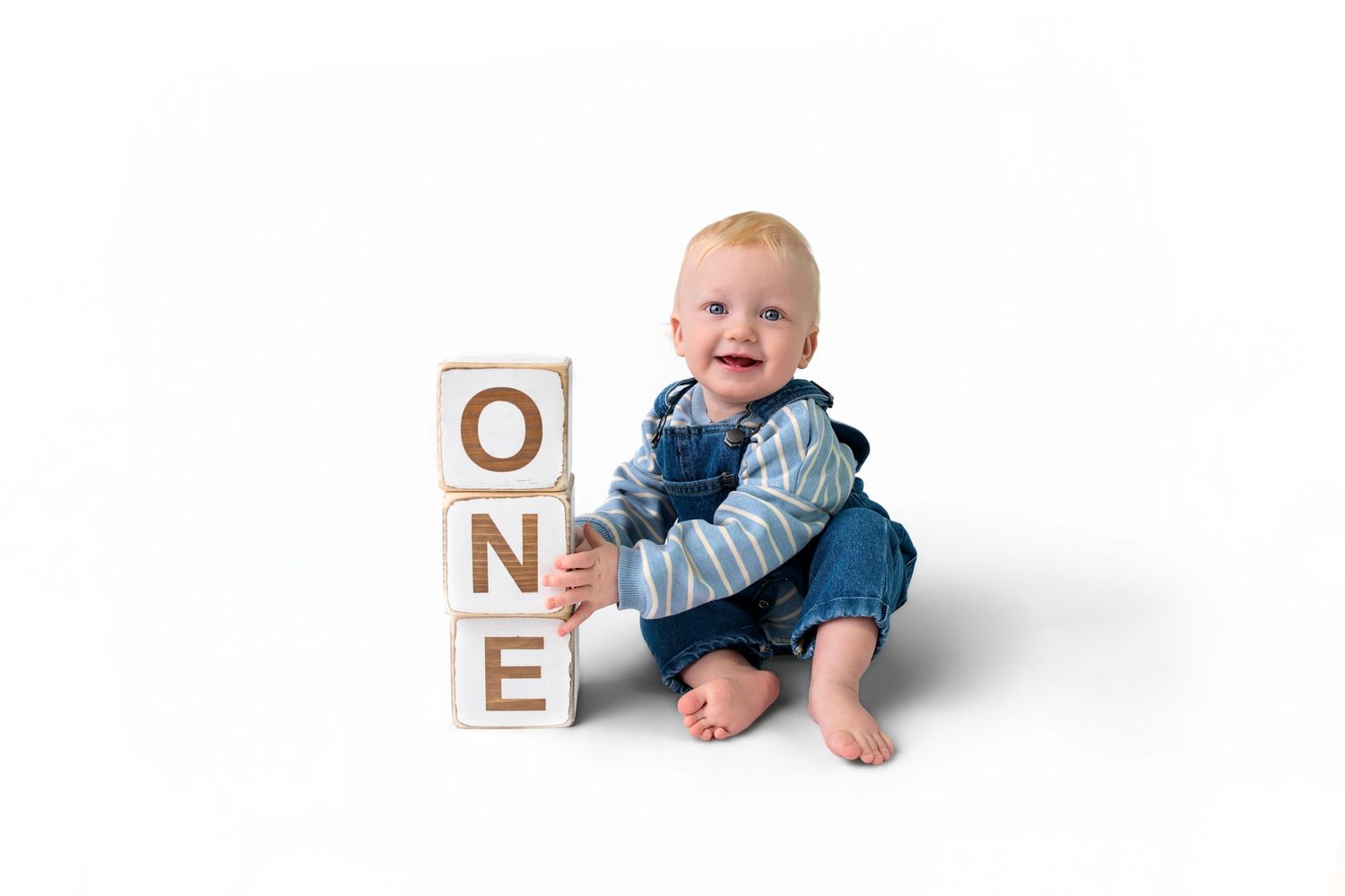 Smiling baby boy sitting next to wooden blocks spelling ONE, celebrating his first birthday milestone.