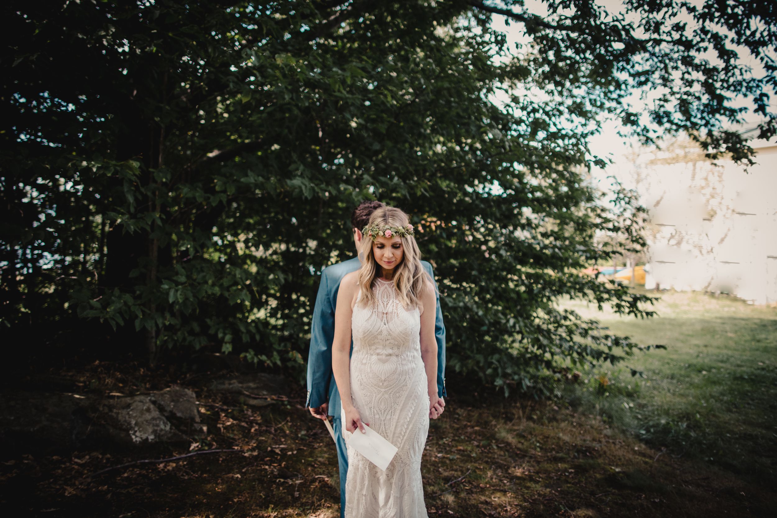 Bride in lace gown with floral crown stands with groom in blue suit under green trees at outdoor wedding.