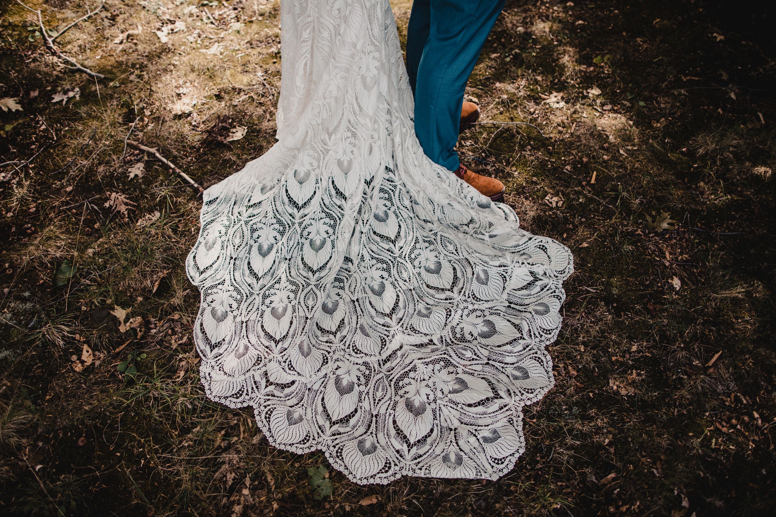 Intricate lace wedding dress train spread on forest ground with peacock feather pattern, bride and groom standing behind.