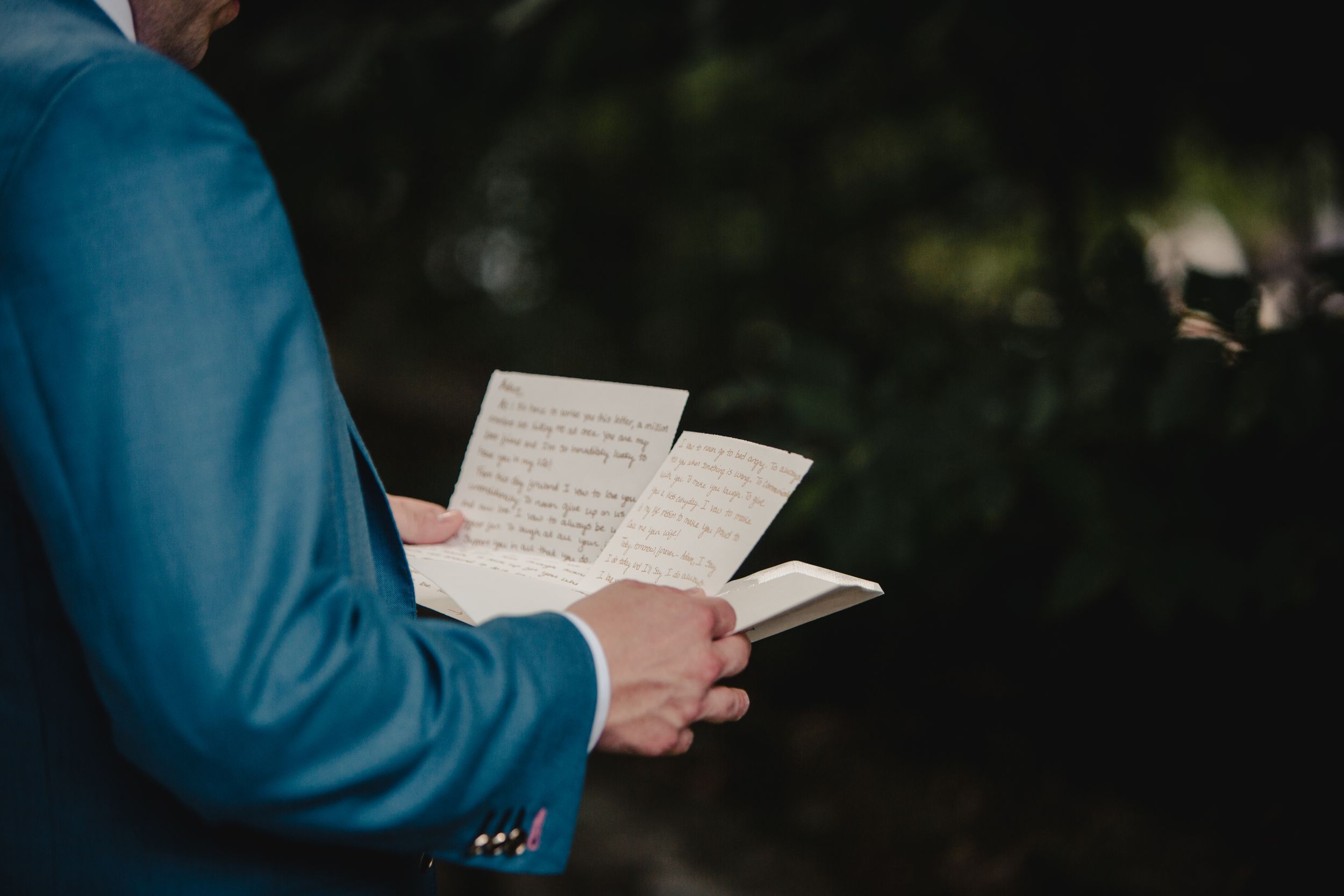 Man in blue suit reading handwritten wedding vows from paper cards outdoors.