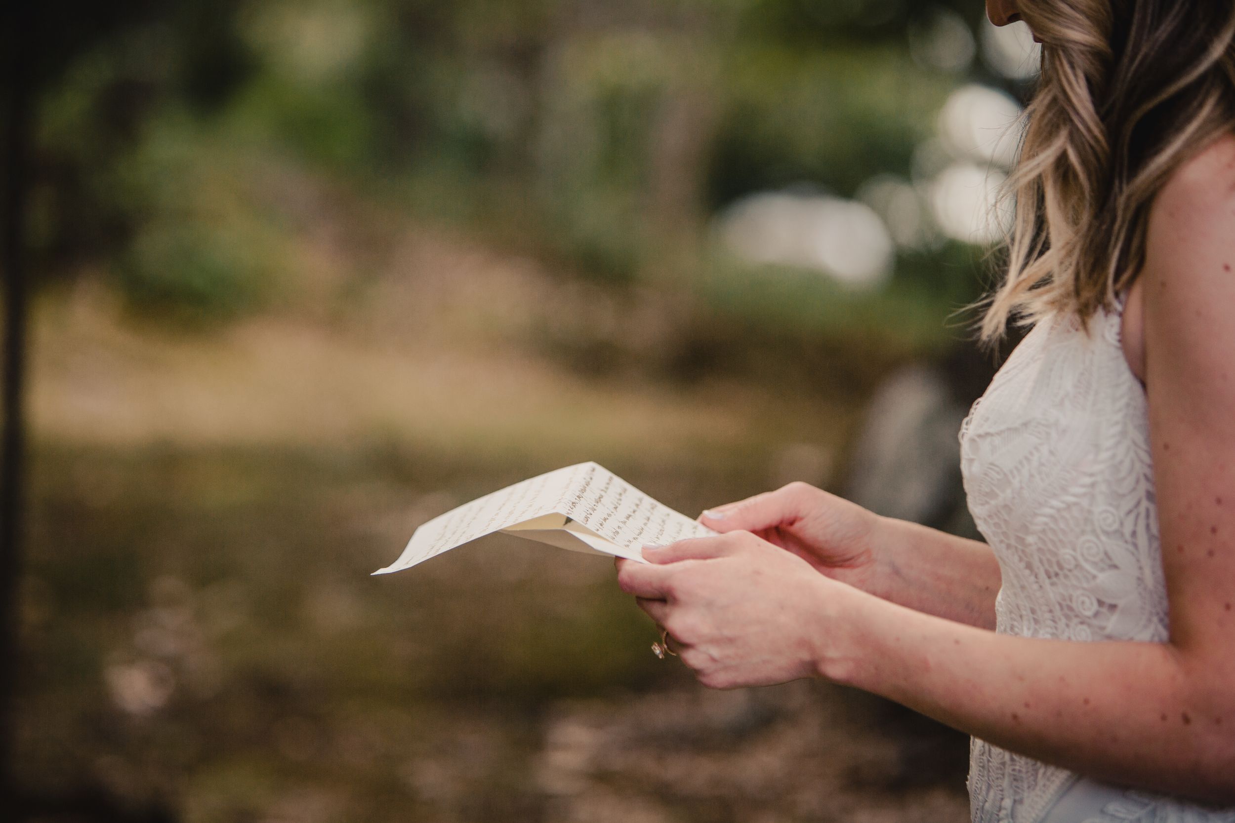 Bride in white lace dress reading handwritten vows on paper outdoors during wedding ceremony.