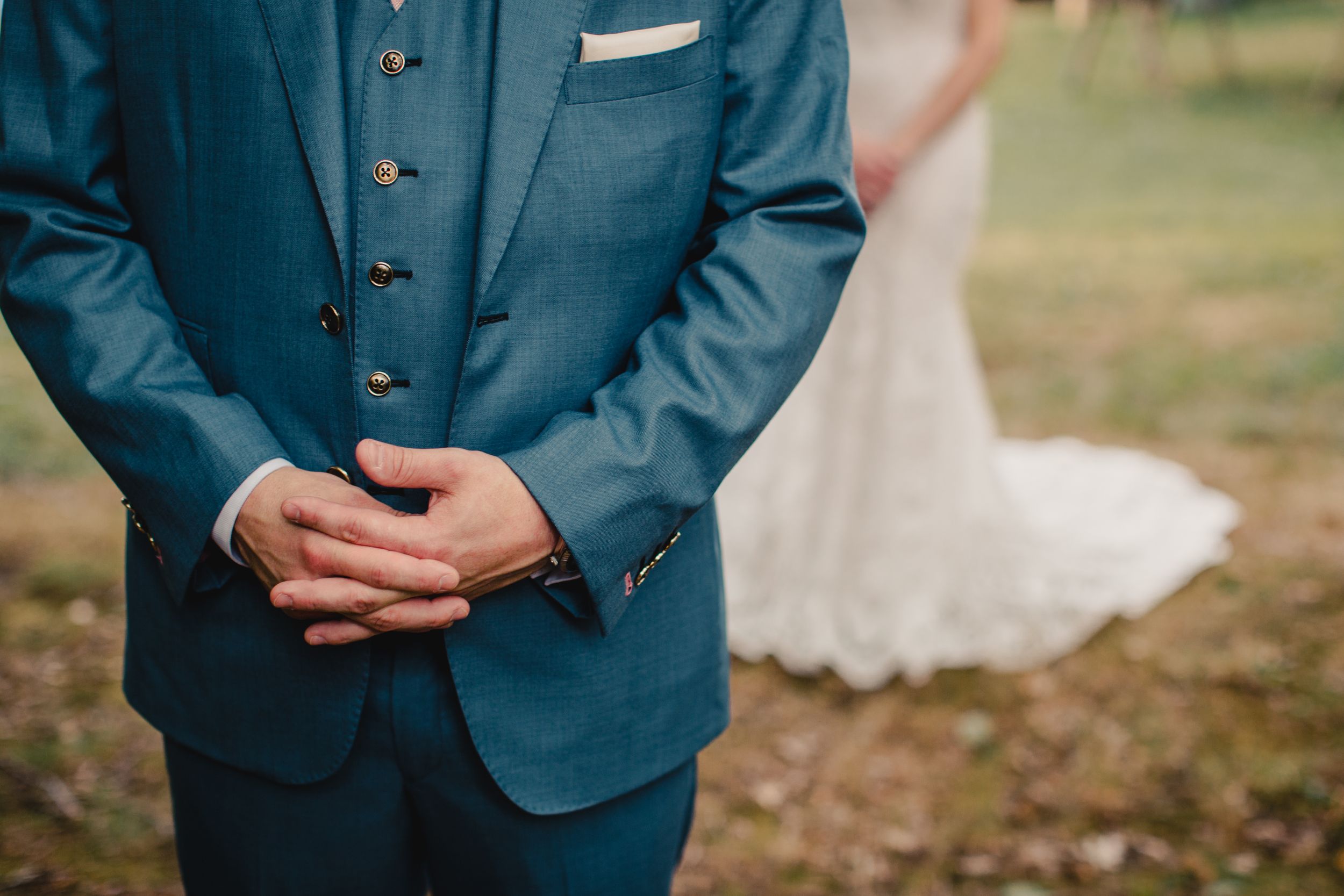 Groom in blue suit clasping hands at outdoor wedding ceremony with bride in lace gown in background.