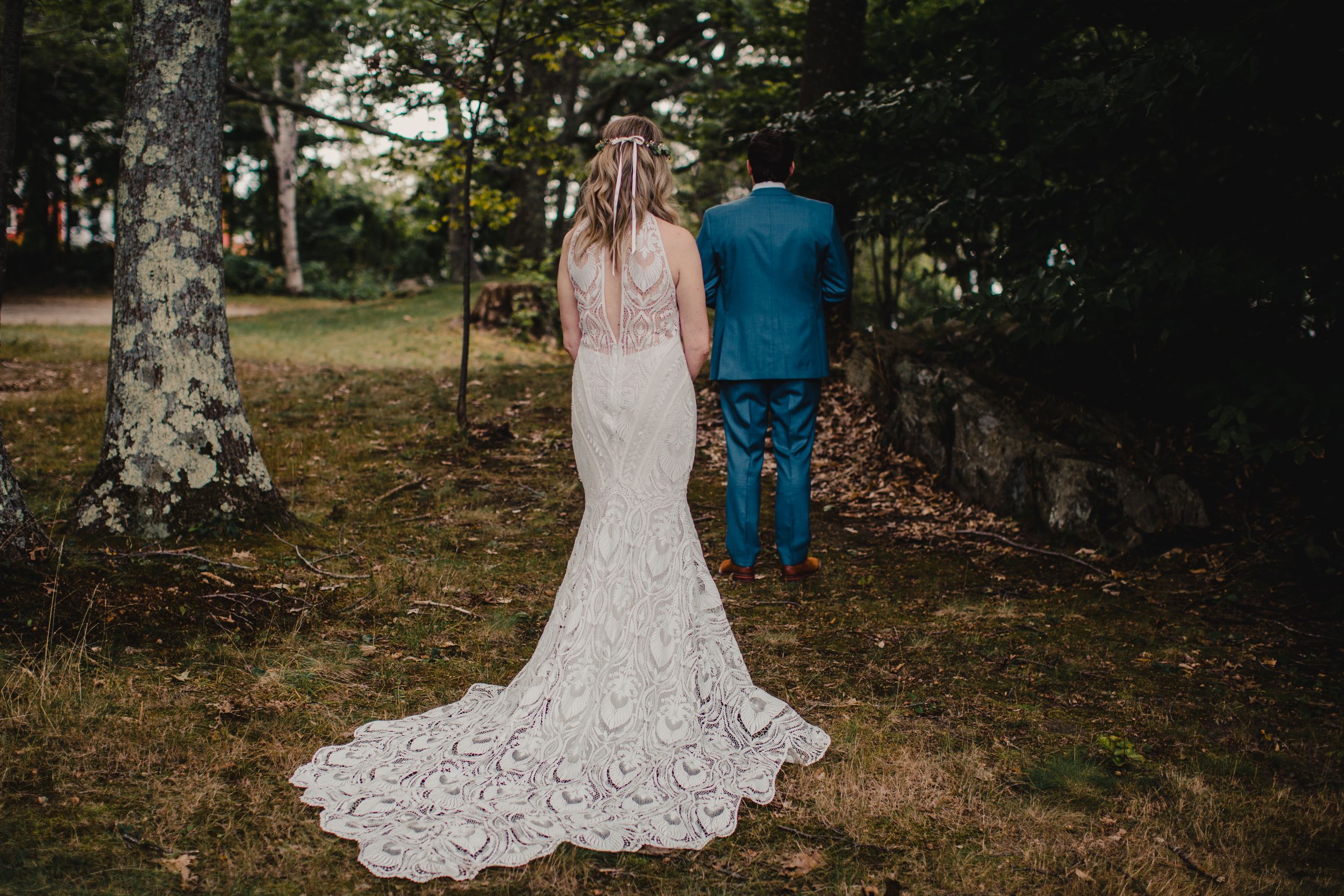 Bride in lace mermaid gown with train stands beside groom in blue suit outdoors in forest setting.