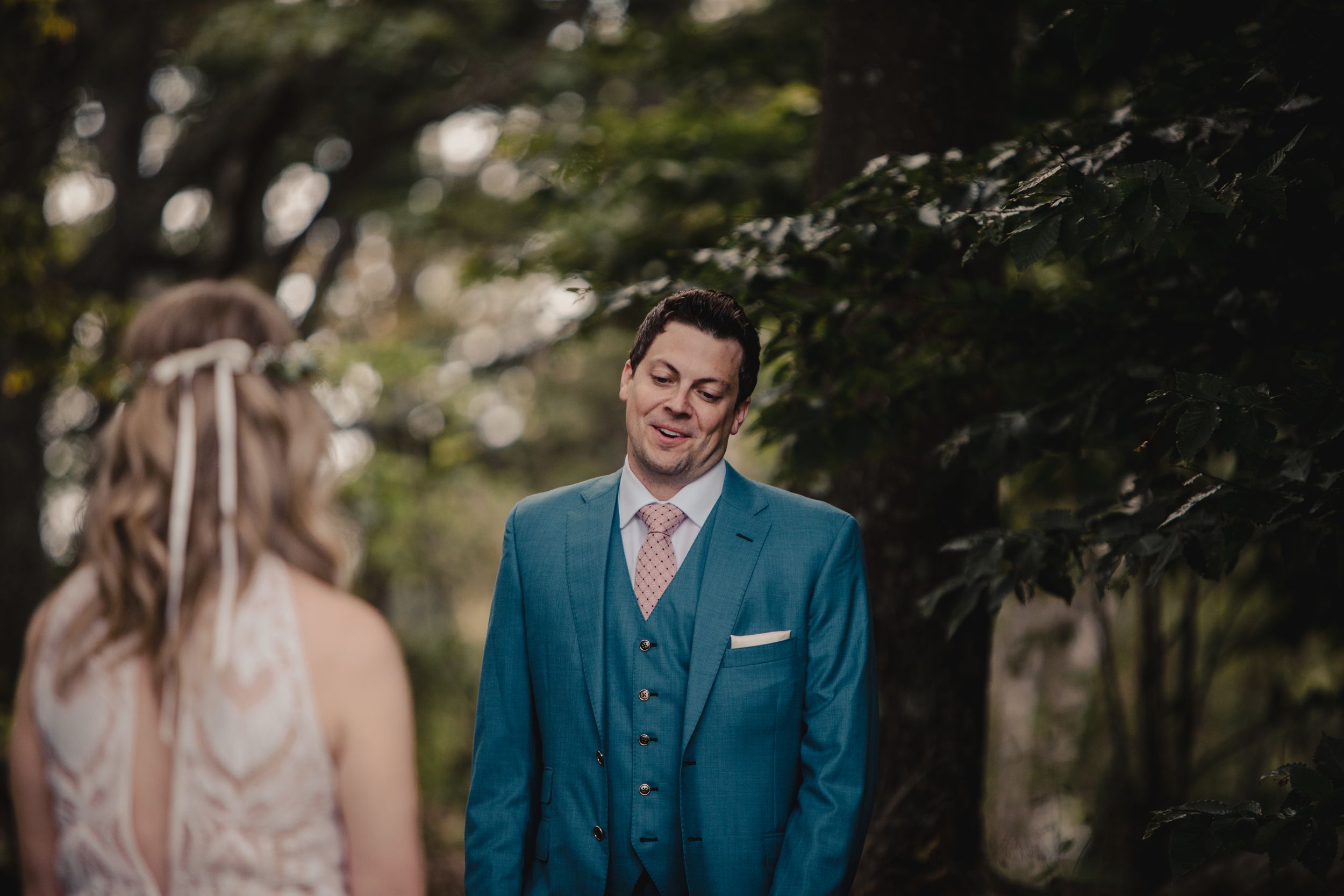 Groom in teal suit smiling at bride during outdoor first look wedding moment among lush green trees.