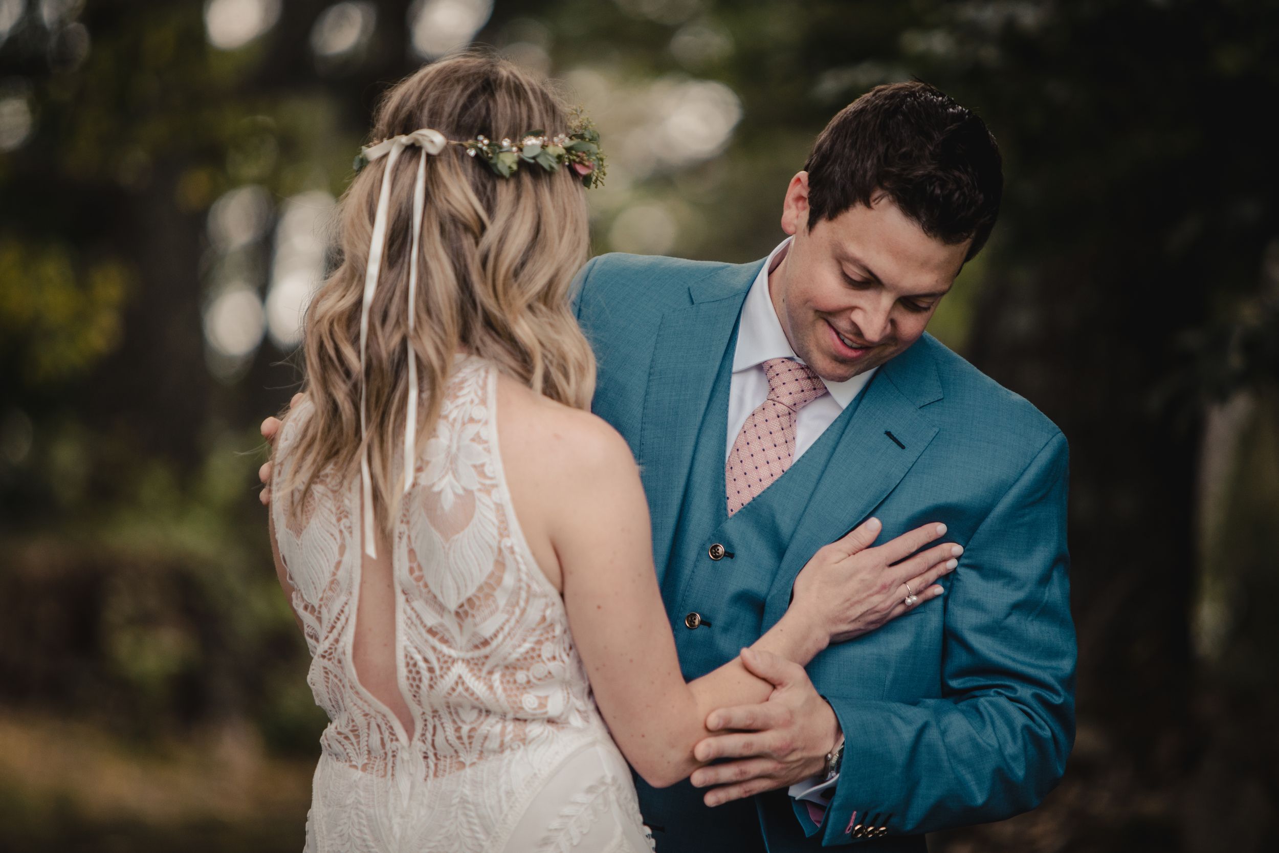 Groom in teal suit smiles as bride in lace wedding dress with floral crown holds his hands outdoors.