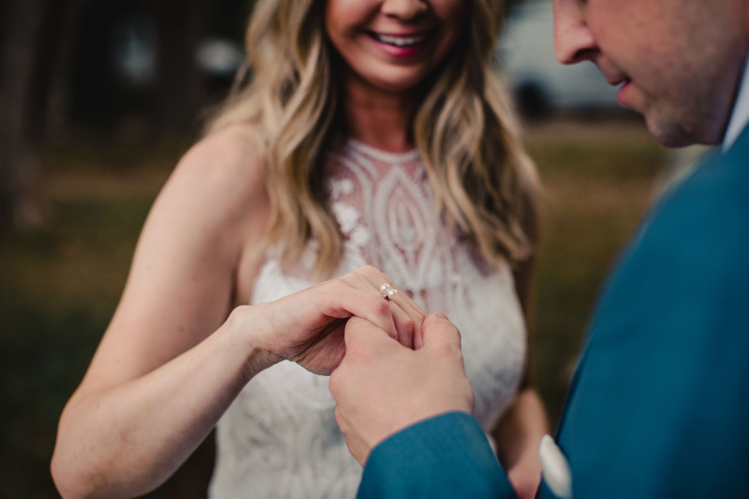 Groom placing wedding ring on bride's finger during outdoor ceremony, bride smiling in lace dress.