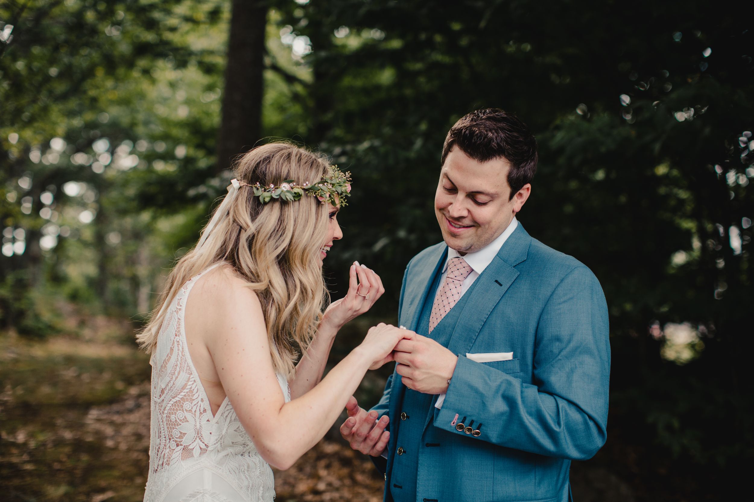 Bride placing wedding ring on groom's finger outdoors in a forest setting, bride wearing floral crown.