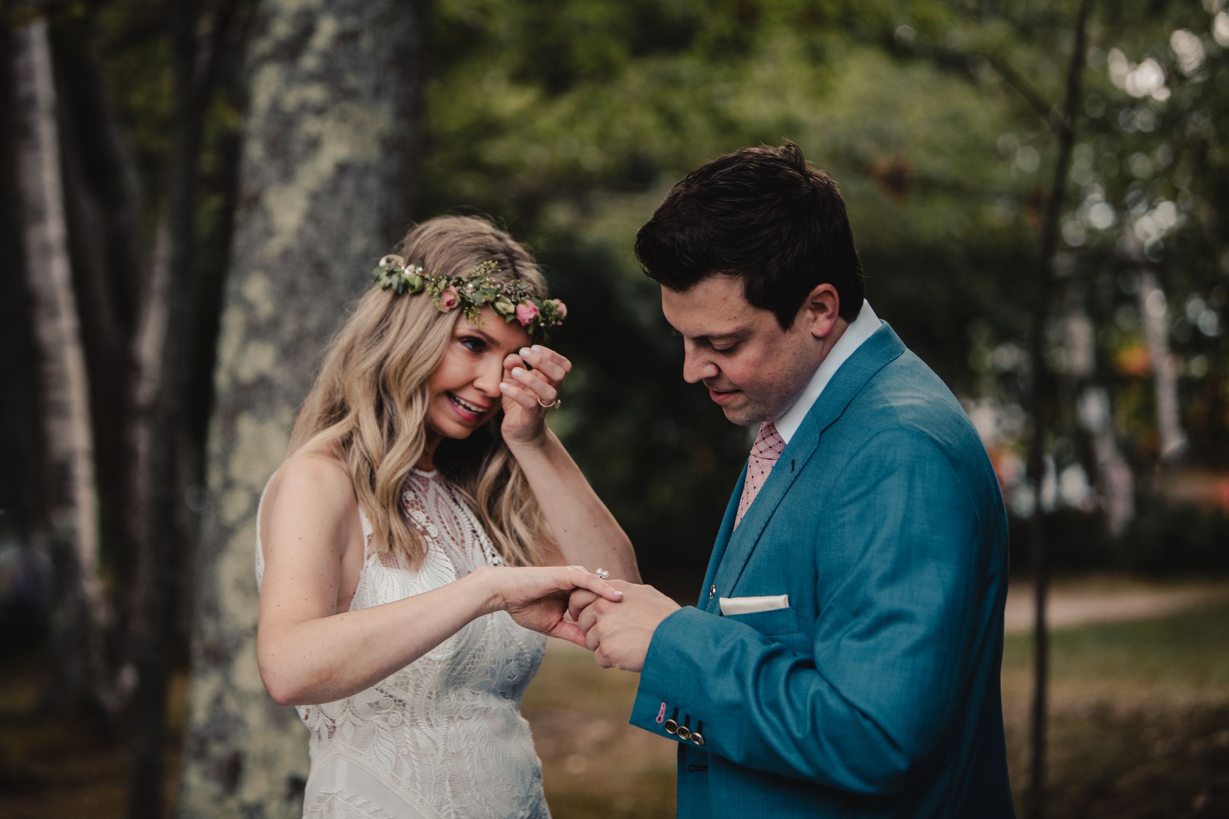 Bride in floral crown and groom in blue suit exchanging rings outdoors during a romantic woodland wedding ceremony.