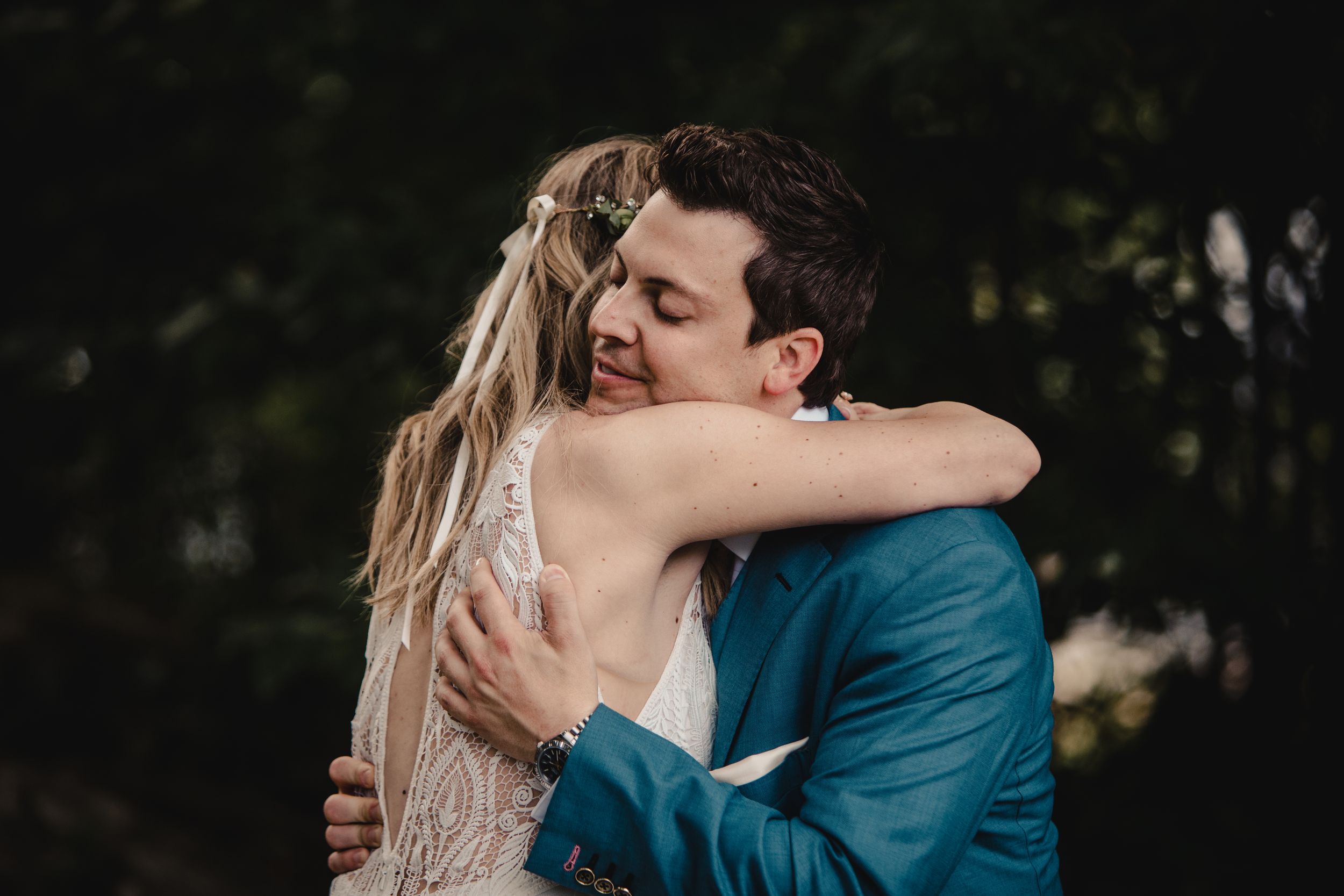 Bride and groom share a heartfelt embrace in forest setting, bride in lace gown, groom in teal suit.