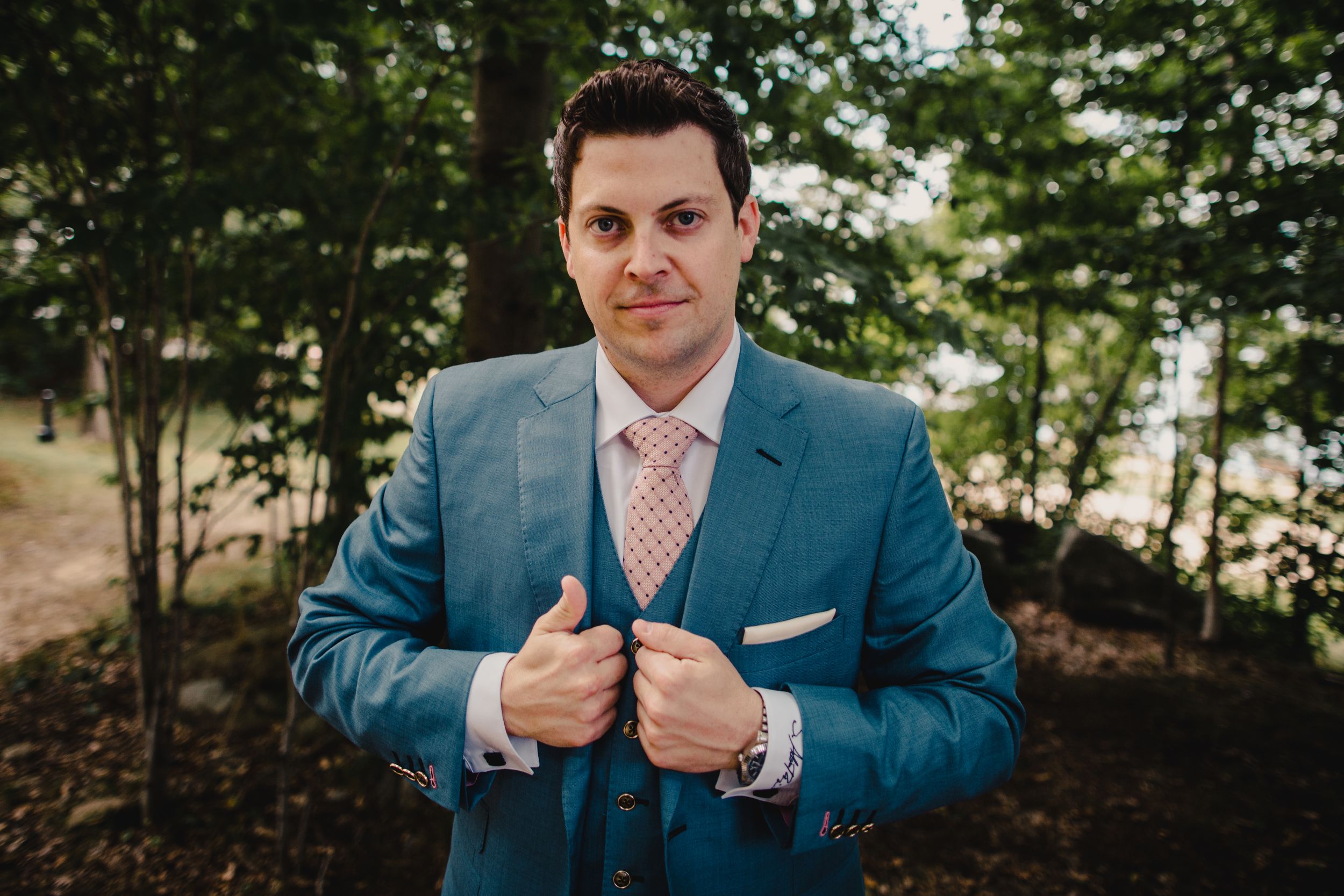 Groom in teal three-piece suit adjusting jacket lapels outdoors with green trees in background.