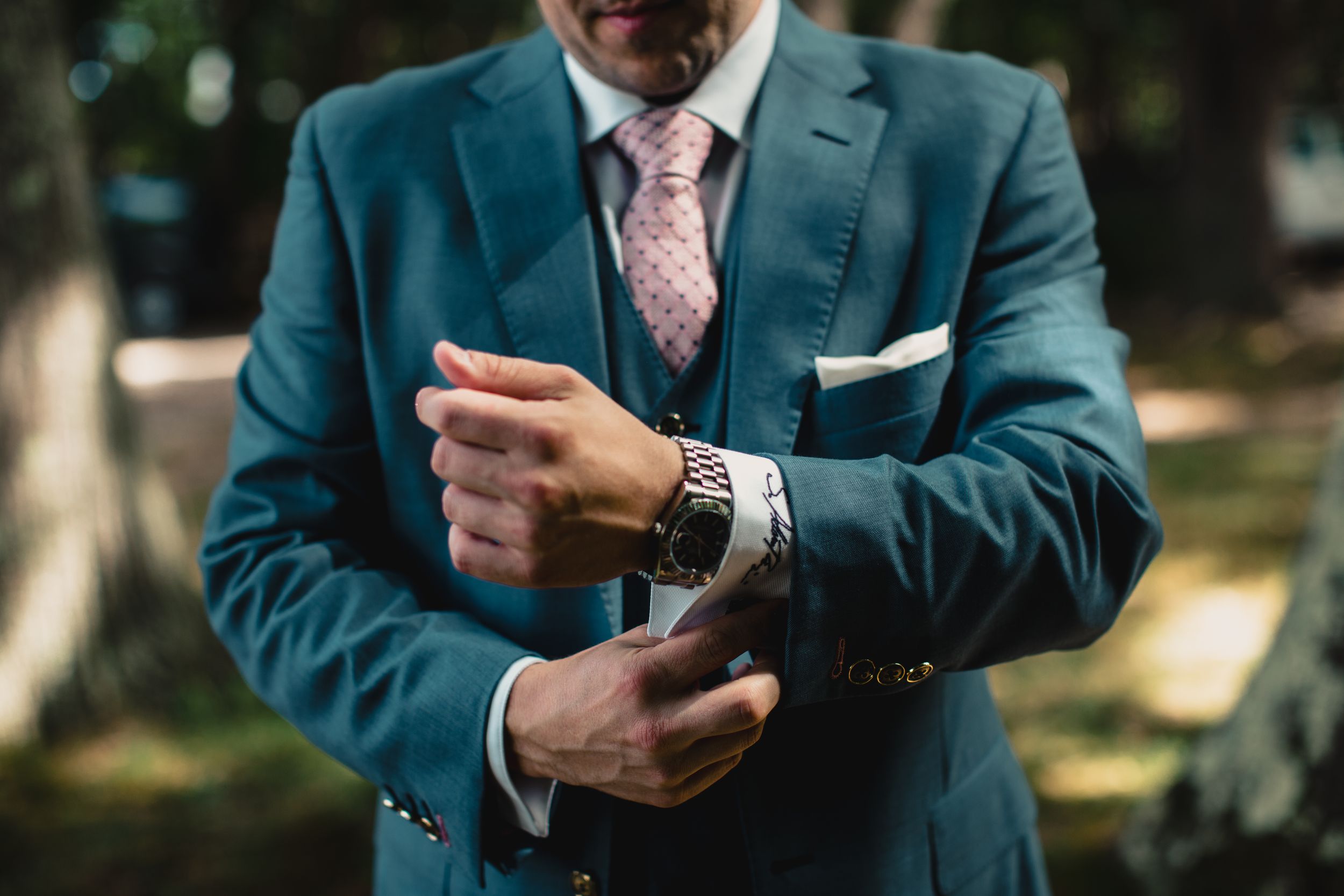 Man in blue suit adjusting watch cufflinks, wearing pink tie and white pocket square outdoors.
