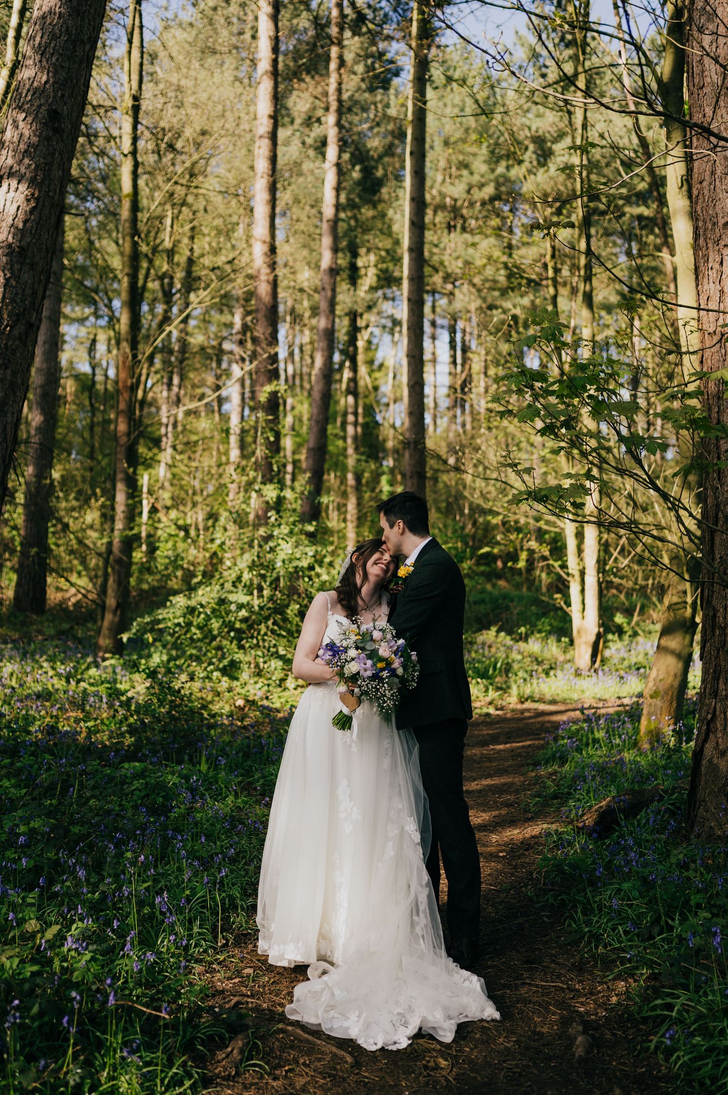 Bride and groom share a romantic moment in a sunlit bluebell woodland, bride holding a pastel bouquet.