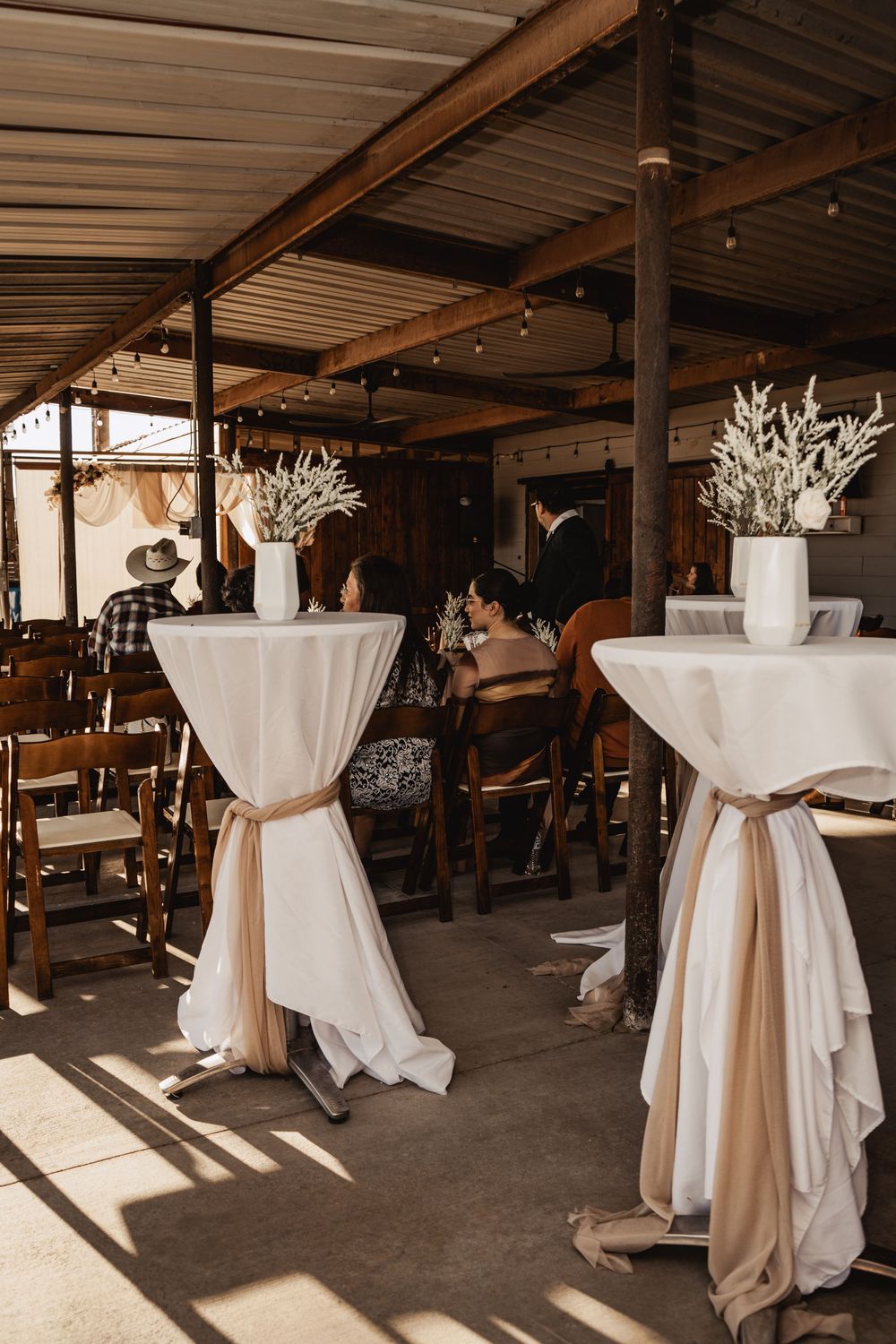 Elegant cocktail tables with white linens and champagne sashes stand in a rustic venue decorated with dried floral arrangements.