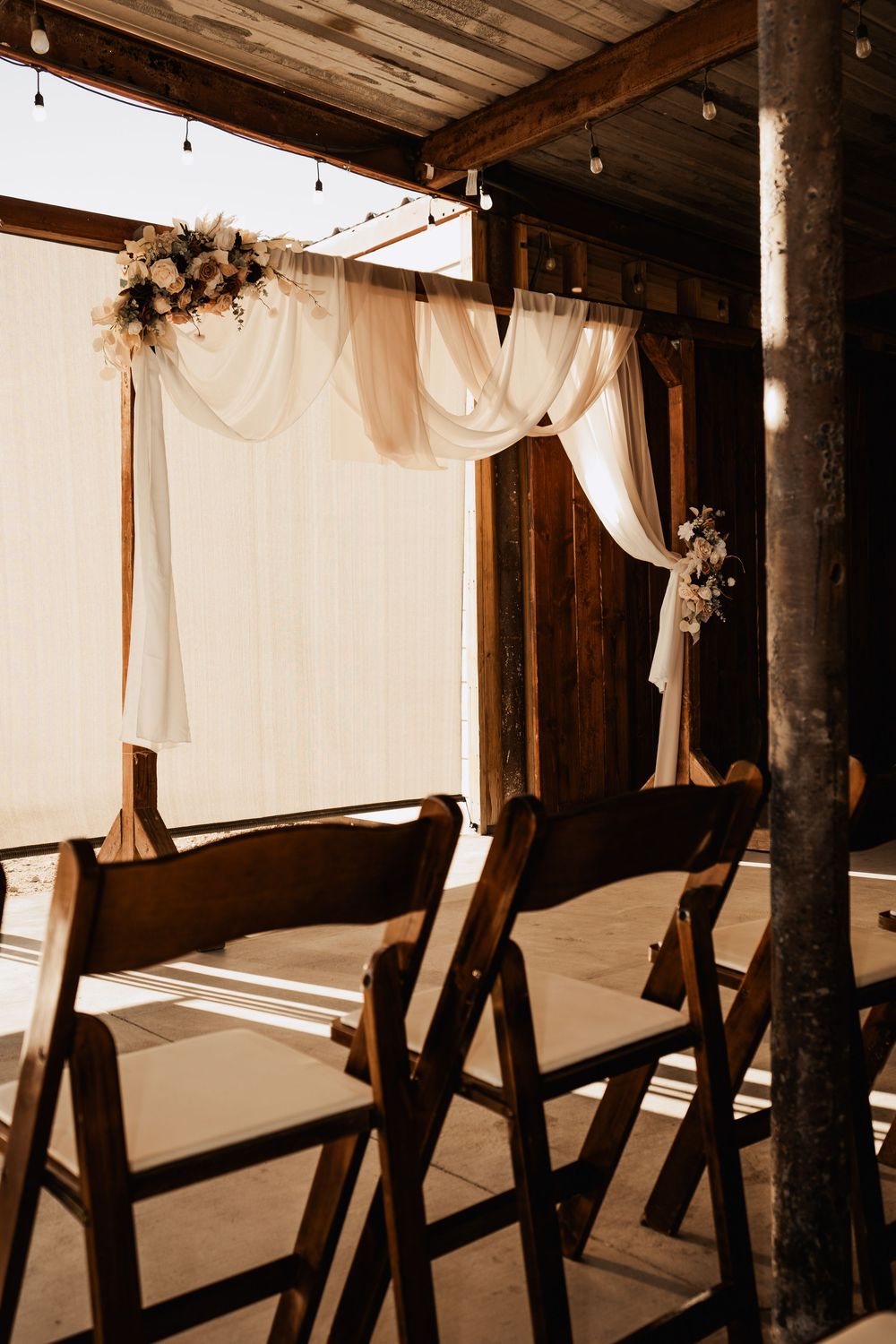 Rustic wedding ceremony setup with wooden folding chairs and flowing white fabric draped from wooden beams with floral accents.