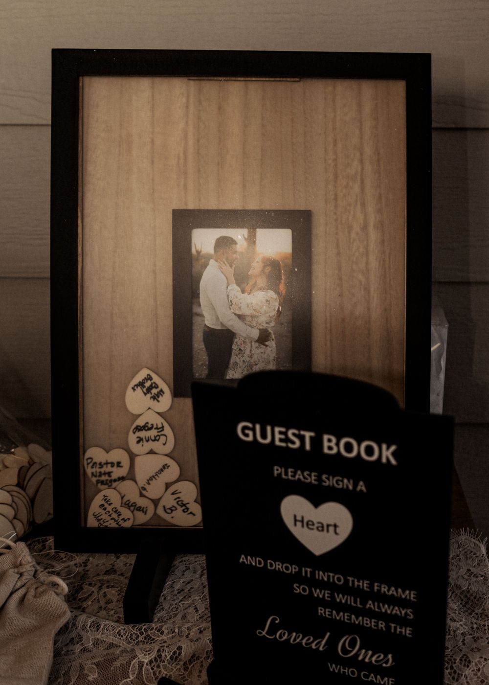 A guest book display with a black frame and wooden background featuring heart-shaped signing cards at a wedding.