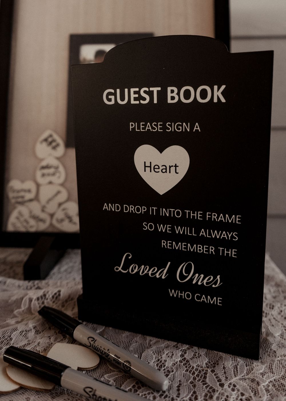 Black guest book sign with heart design asking guests to sign and drop hearts into a frame to remember loved ones.
