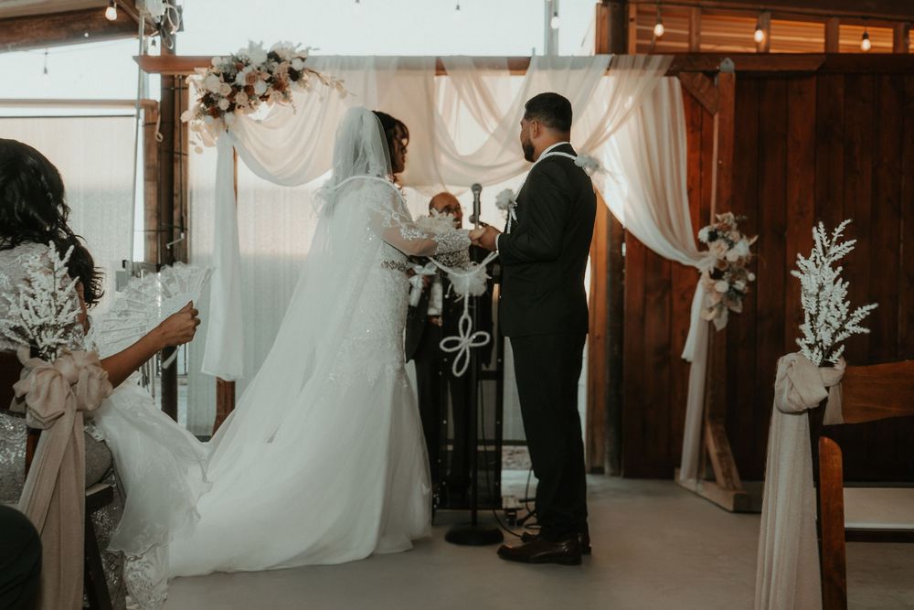 A couple stands at a wedding ceremony altar with white drapes and floral decorations in a rustic indoor venue.