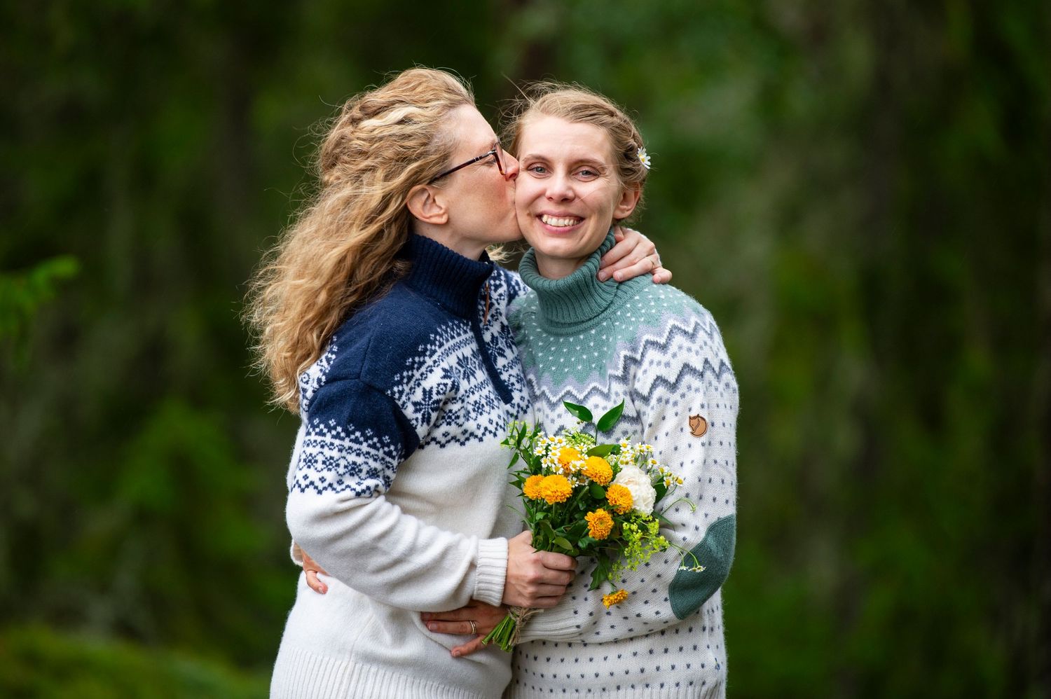 Two people in cozy Nordic sweaters share a sweet moment outdoors while holding a bouquet of yellow flowers.
