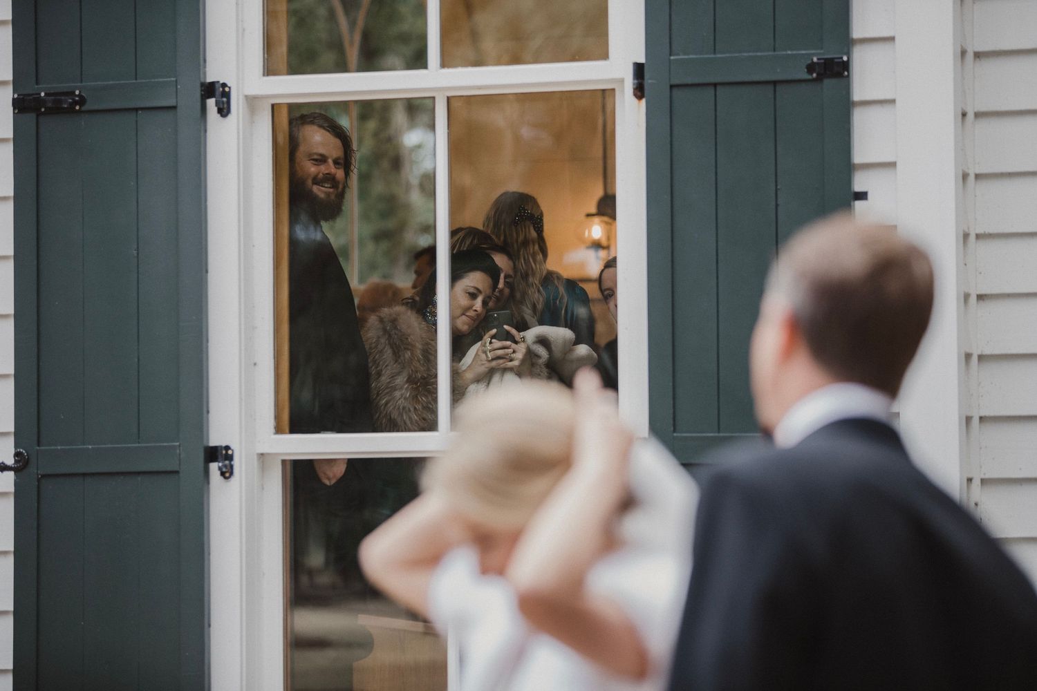 People looking through a window with green shutters during what appears to be a wedding moment.