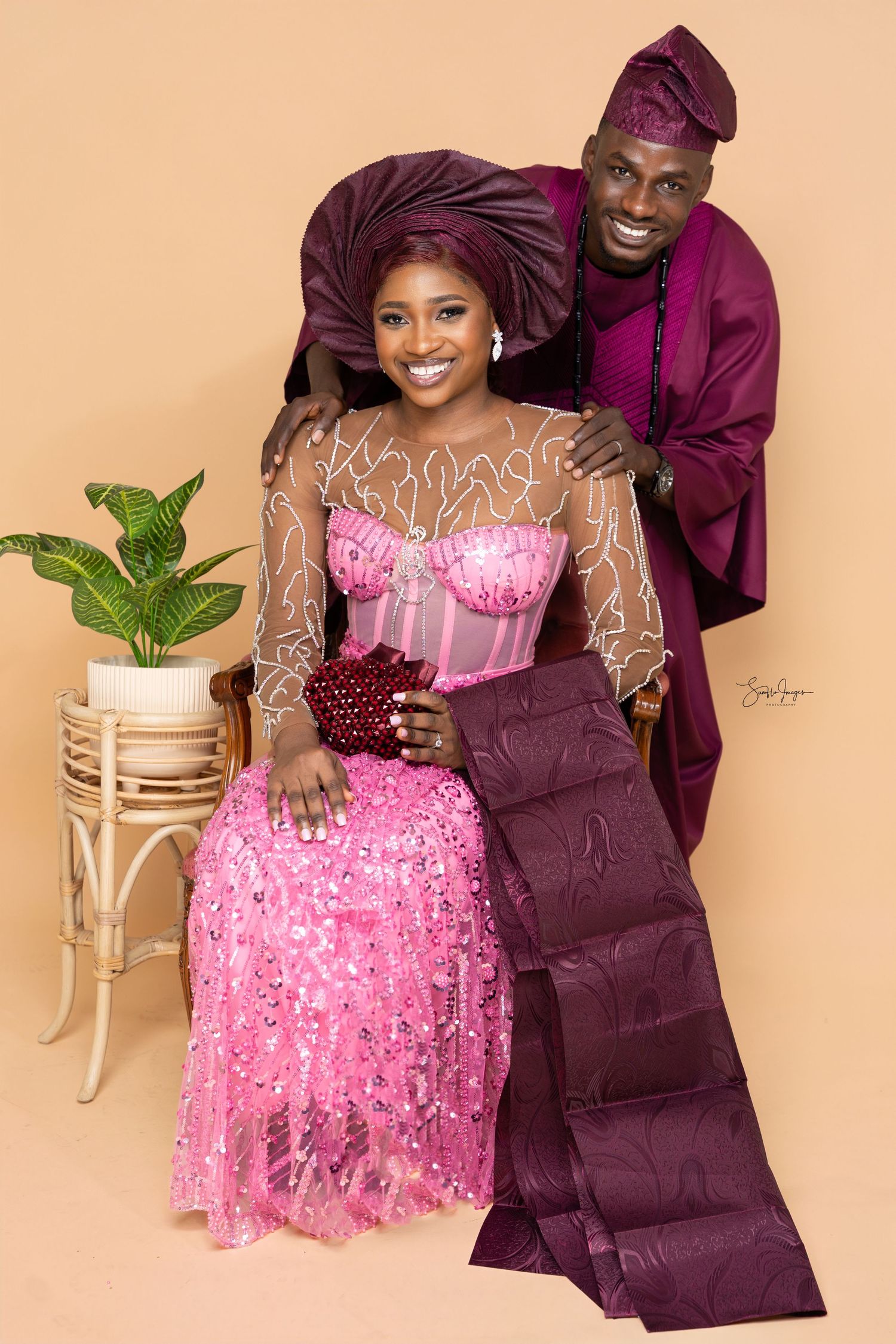 A couple poses in coordinated burgundy and pink formal attire against a beige backdrop with decorative plant accents.