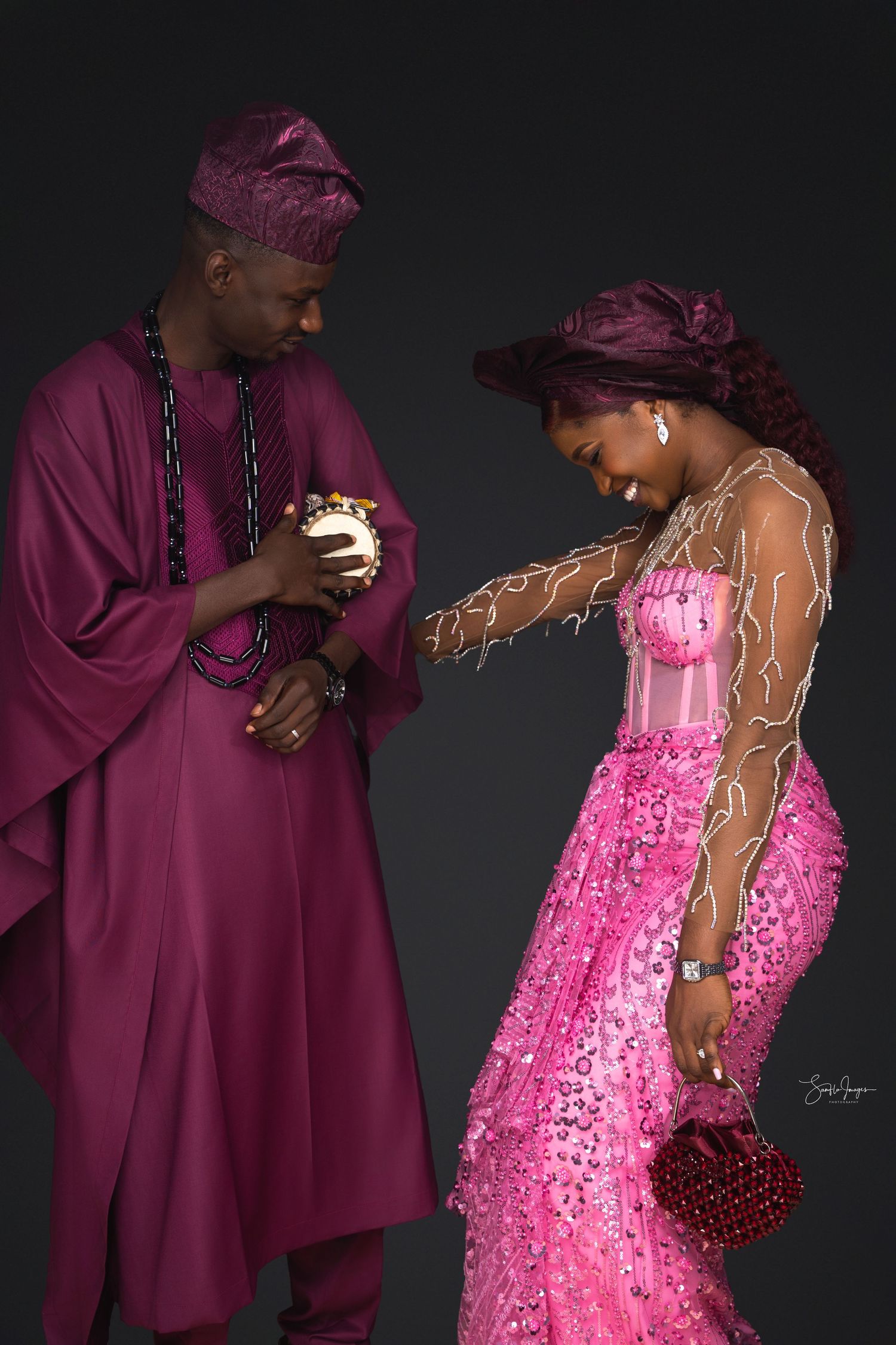 A couple poses together in contrasting burgundy suit and sparkly pink dress against a dark background.