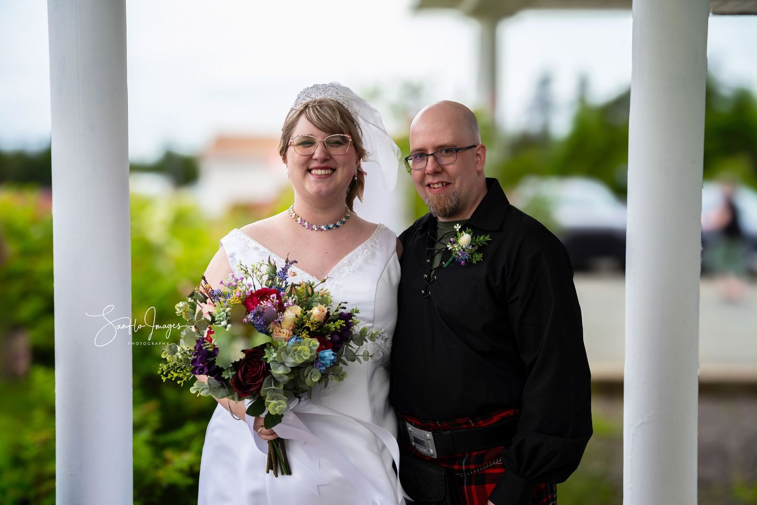 A wedding couple poses together between white columns on their special day.