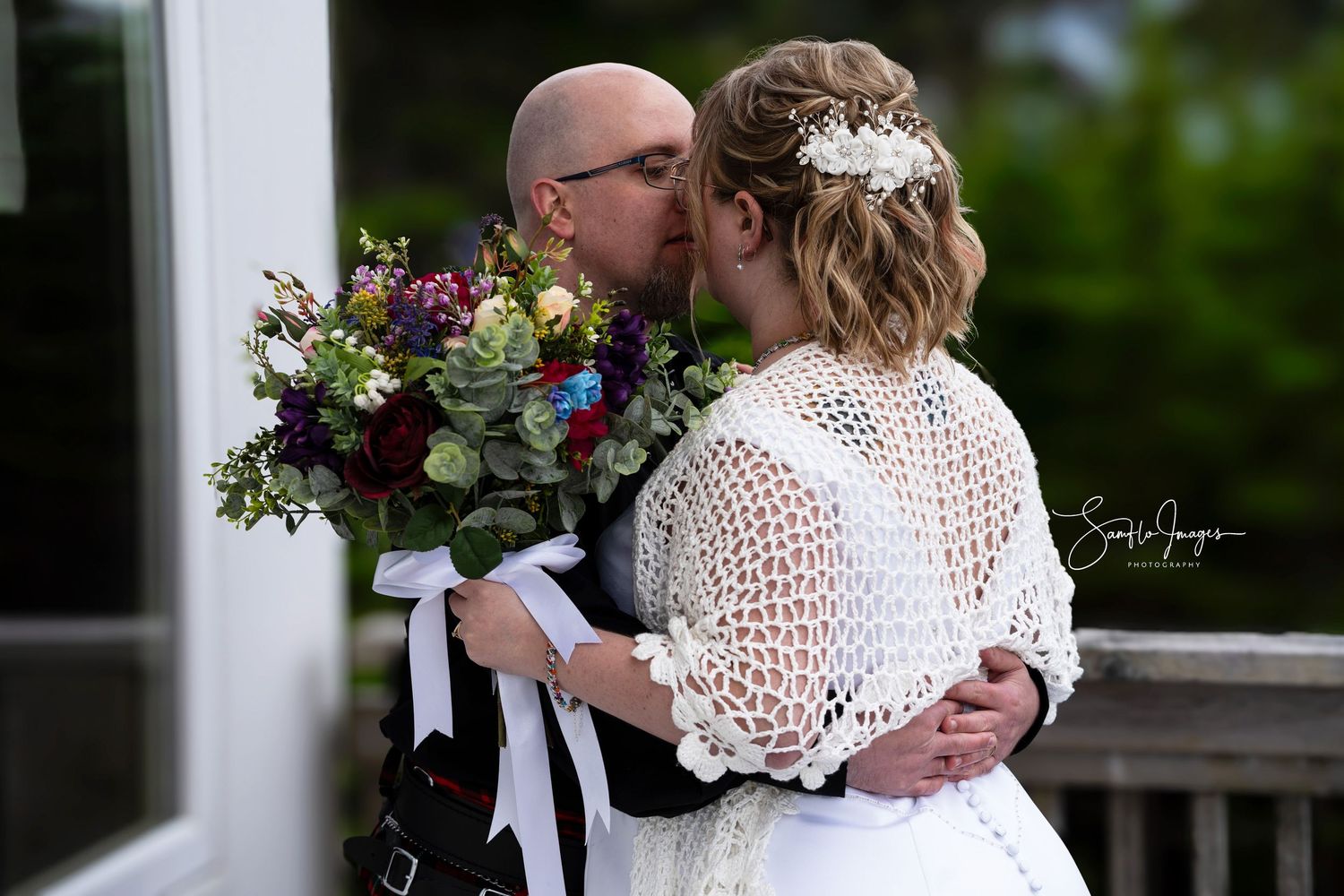 A couple in white attire embraces during an outdoor wedding celebration with floral arrangements and greenery.