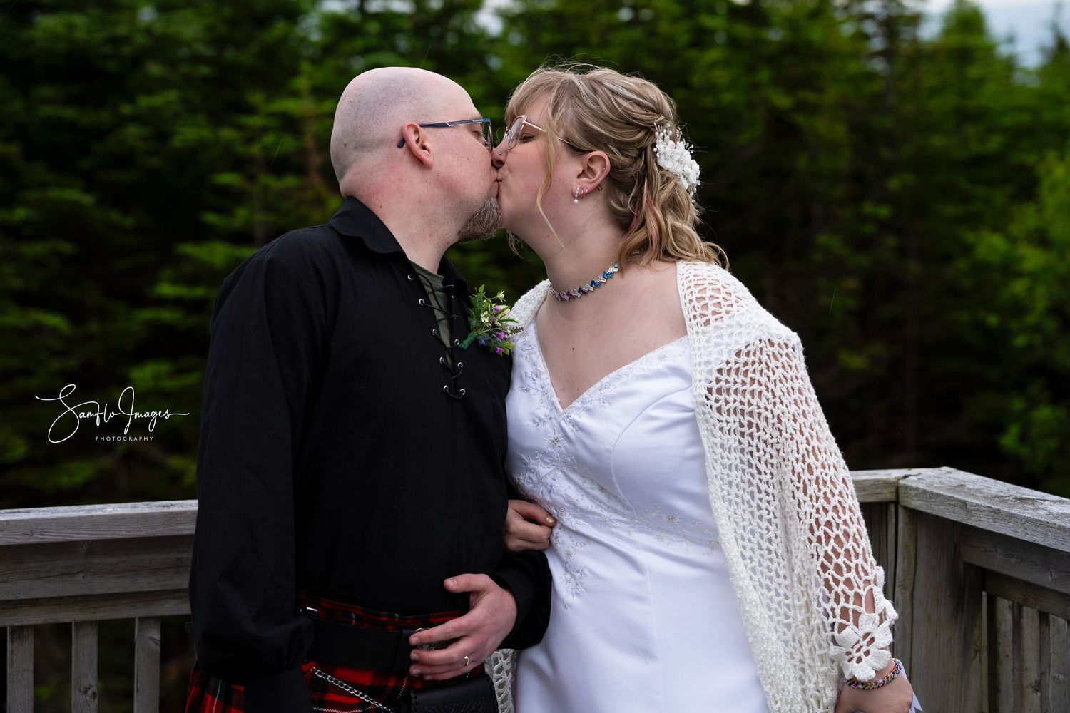 A couple shares an intimate moment on a wooden deck surrounded by evergreen trees in a romantic outdoor setting.