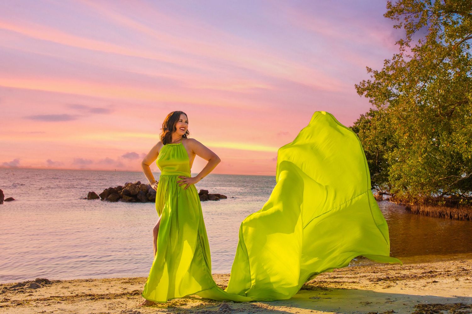 A flowing neon yellow dress billows in the wind against a pink sunset sky on a tropical beach.