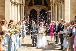 Newlyweds walk through a shower of confetti outside a Gothic cathedral surrounded by guests.