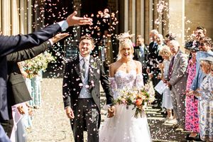 Smiling newlyweds exit a grand cathedral as guests throw pink and white confetti in celebration.