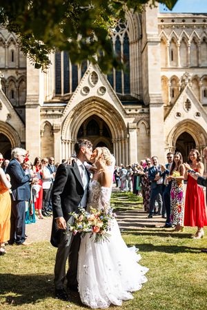 A newlywed couple shares a kiss outside a stunning Gothic cathedral while guests throw confetti in celebration.