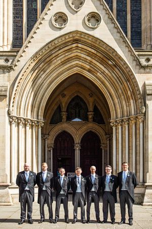 Seven groomsmen in morning suits pose beneath the Gothic arched entrance of a historic cathedral.