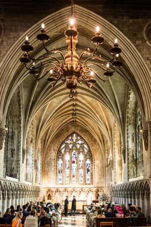 Gothic cathedral interior with vaulted ceiling, ornate chandelier, and stained glass window during wedding ceremony.