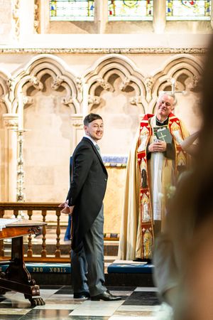 Smiling groom in black suit standing at church altar with robed officiant during wedding ceremony.