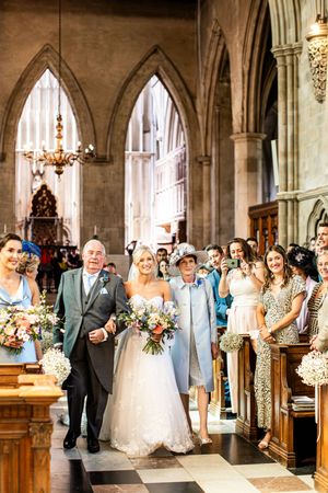 Bride in strapless lace gown walks down church aisle with parents as wedding guests look on joyfully.