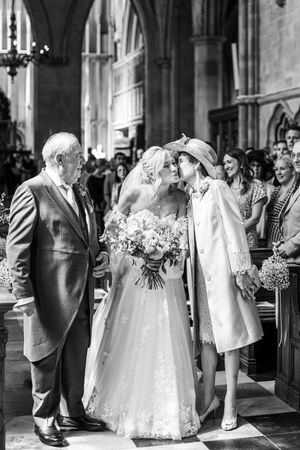 Black and white photo of bride in lace gown kissing mother while father stands beside her at church aisle.