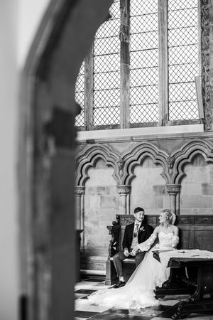 Black and white photo of bride and groom seated together beneath a grand arched church window.