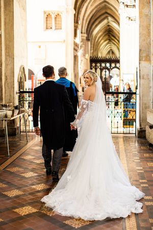 Bride in a flowing lace gown and groom walk hand in hand through a stunning cathedral aisle.