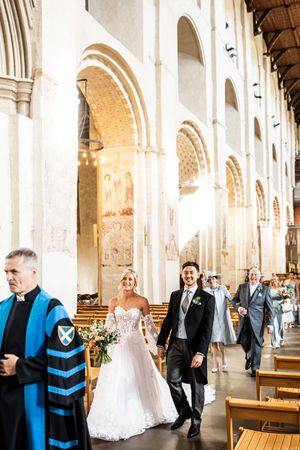 Newlyweds walk joyfully down the aisle of a grand cathedral accompanied by a minister in blue robes.