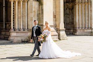 Bride in a flowing white gown and groom walk hand in hand outside a grand stone cathedral.