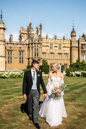 Bride and groom walking hand in hand in front of a grand historic English manor house.
