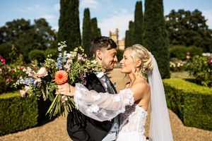 Bride and groom laughing together in formal garden holding colorful wedding bouquet with topiary hedges and mansion backdrop.
