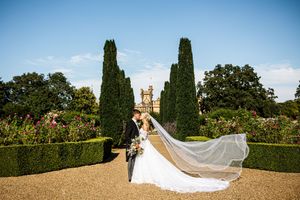Bride and groom kissing in formal garden with flowing veil billowing in wind, tall cypress trees and stately home behind.