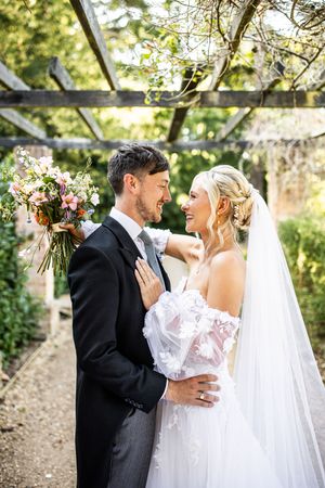 Smiling bride and groom embracing under rustic pergola, bride in off-shoulder lace gown holding pink floral bouquet.