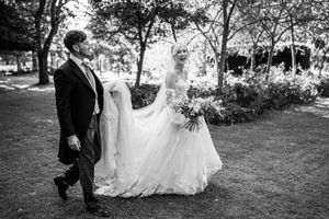 Black and white photo of groom carrying bride's train as they walk through sunlit garden grounds together.