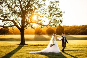 Bride and groom holding hands walking across golden sunlit meadow at sunset with large oak tree in background.