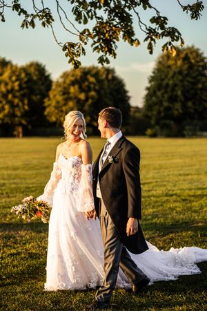 Bride and groom walking hand in hand through open parkland at golden hour, bride in lace off-shoulder wedding gown.