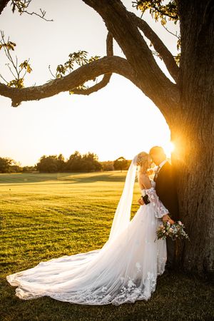 Bride and groom kissing beneath large oak tree at sunset, bride's cathedral veil flowing across lush green lawn.