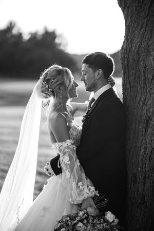 Black and white romantic portrait of bride and groom gazing at each other leaning against large tree trunk.